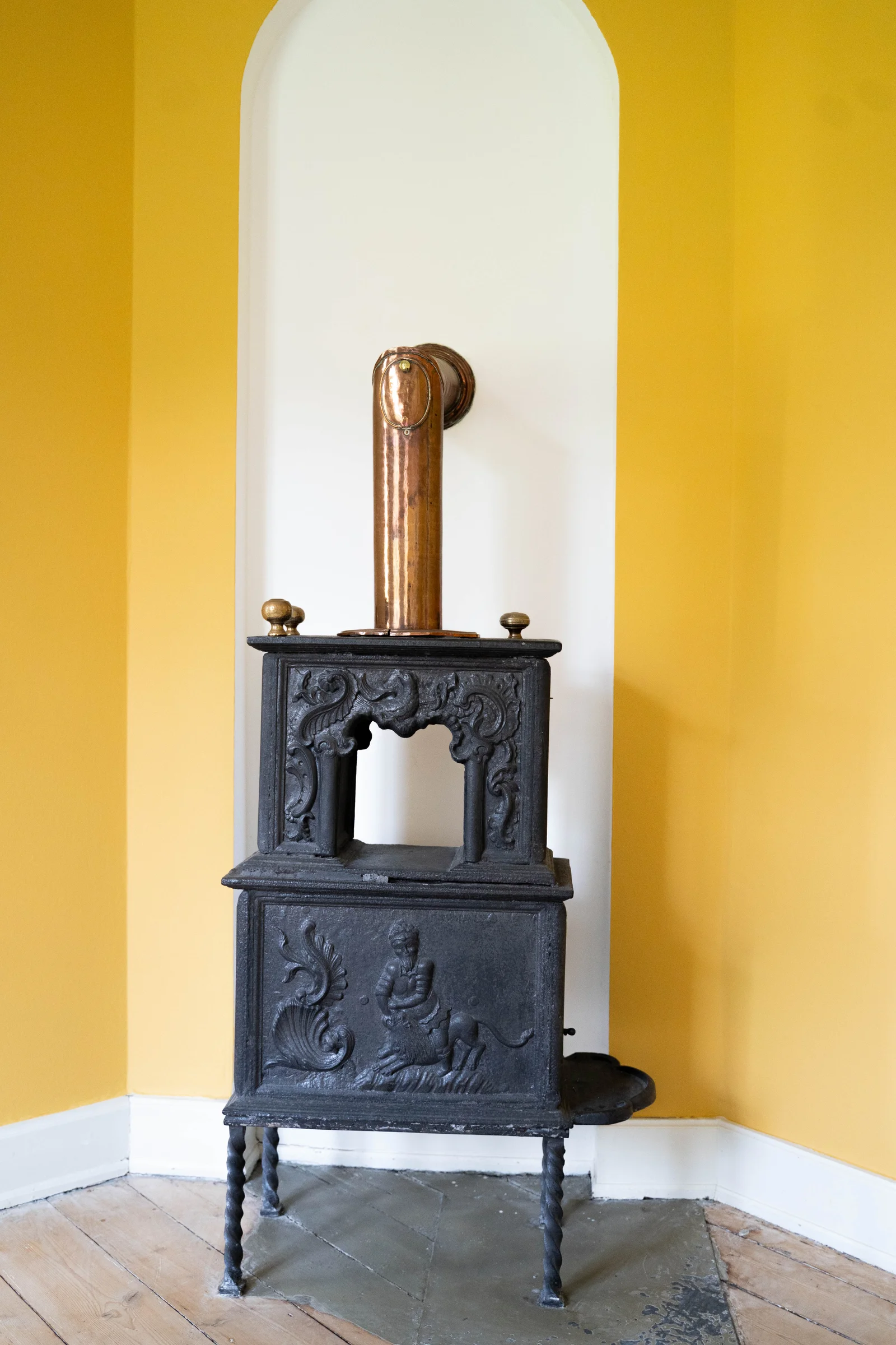 Ornamentierter Gusseisenofen mit Kupferrohr in einer gelbwandigen Nische im Gutshaus Agerup