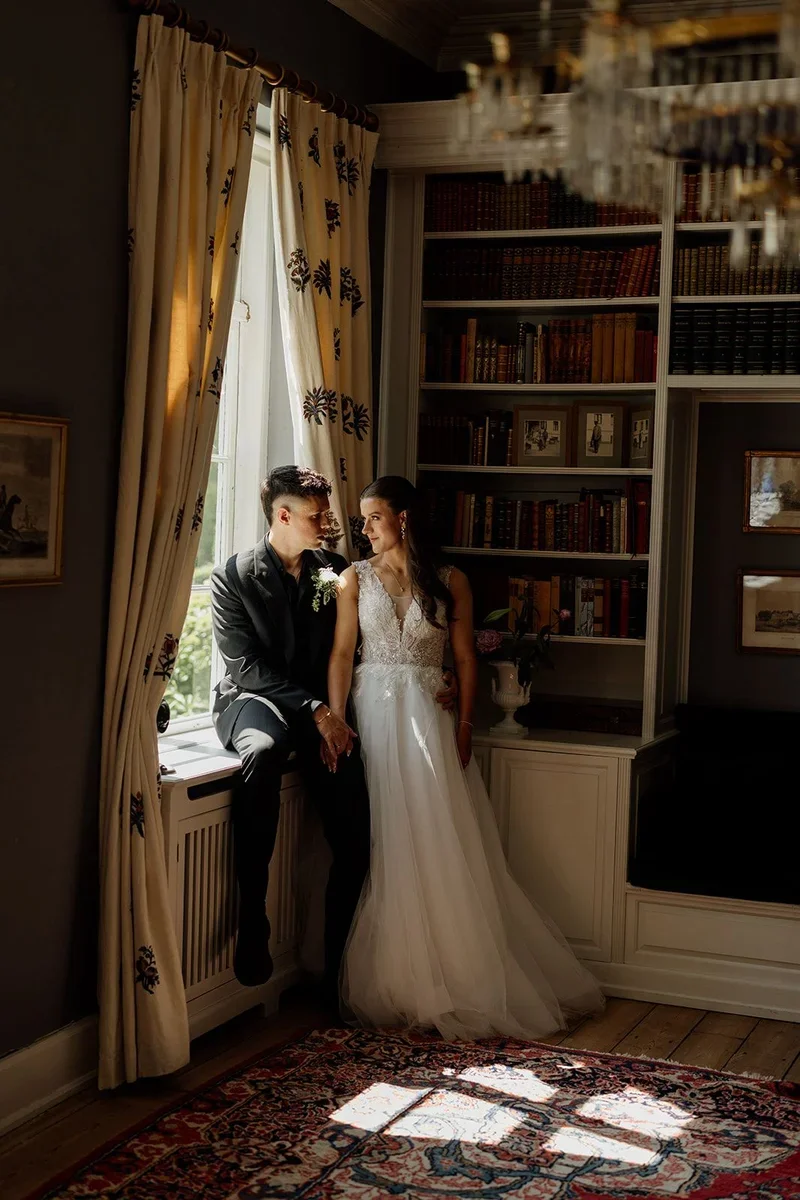 Brudepar ved vinduet i biblioteket på Agerup Gods med antikke bøger og lysekrone
